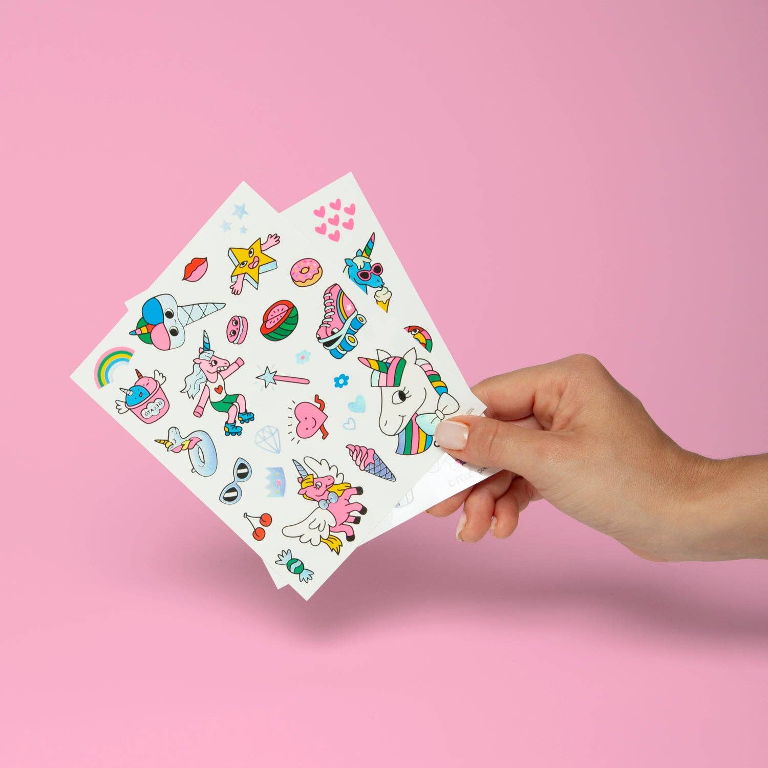 Hand holding two sheets of colorful temporary tattoos against a pink background