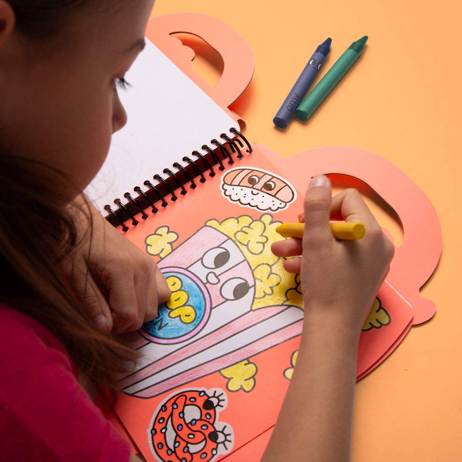 Child coloring a cartoon character in a spiral-bound coloring book with crayons on an orange background.
