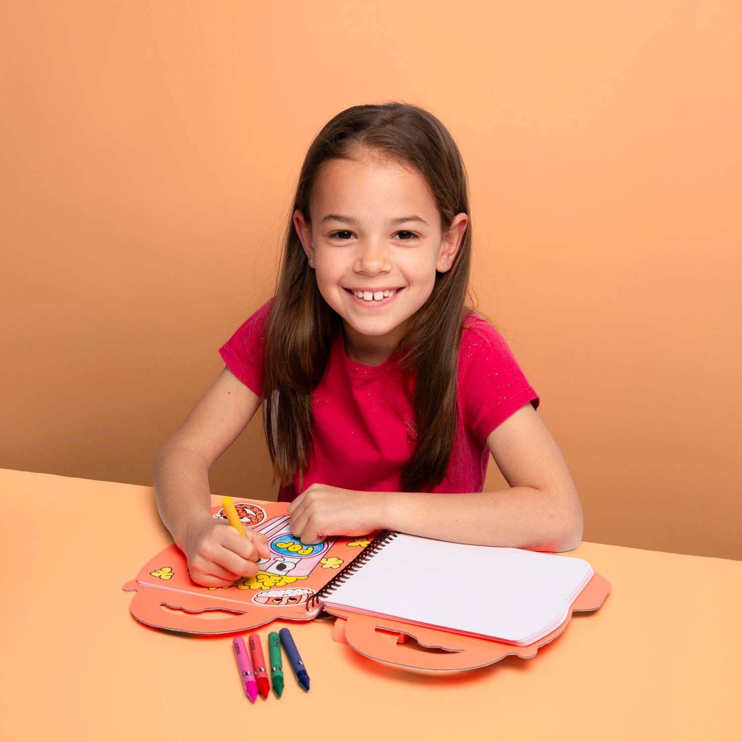 Young girl sitting at a table with a coloring book and markers, smiling against an orange background