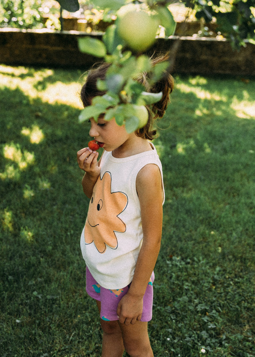 A young kid wearing pink shorts with green designs and a white tanktop with smiling cloud print.